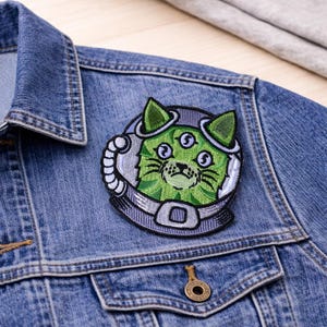 May include: Embroidered patch featuring a green cat with three eyes wearing a space helmet. The patch is attached to a denim jacket. The cat's face is detailed with black outlines and the helmet is gray and white.