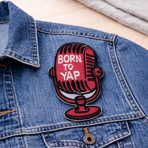 May include: A red and black microphone patch with the words "BORN TO YAP" in white lettering. The patch is affixed to a denim jacket. The microphone design features a retro style, with a stand and a detailed grill.