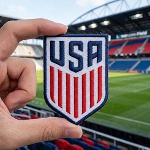 May include: A shield-shaped embroidered patch featuring the letters USA in dark blue at the top, positioned above seven vertical red stripes on a white background, all bordered by a thick dark blue stitched edge on a white fabric base.