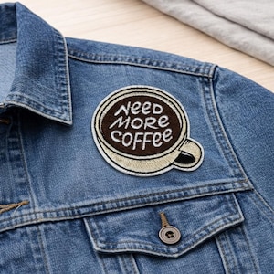 May include: A denim jacket with a patch that reads "Need More Coffee" in a coffee cup design. The patch is embroidered with brown and beige thread. The jacket has a buttoned pocket and a button closure.