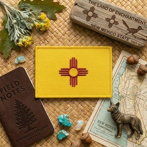 May include: A yellow patch with the New Mexico state flag, featuring a red Zia symbol. The image includes a wooden sign with text, a map, a bronze coyote figurine, a notebook, and turquoise stones. The words "FIELD NOTES" are visible.