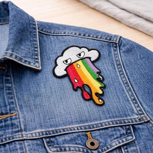 May include: A colorful embroidered patch featuring a white cloud with a grumpy face, spewing a rainbow. The patch is sewn onto a blue denim jacket. The rainbow has red, orange, yellow, green, and purple colors.