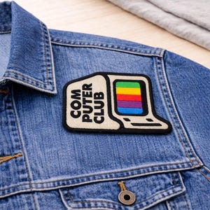 May include: A denim jacket with a patch that reads "COMPUTER CLUB." The patch features a retro computer design with a rainbow-colored screen. The jacket is a light blue wash with visible stitching and a button closure.