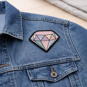 May include: Embroidered diamond patch with pastel pink, purple, yellow, and blue segments, outlined in black. The patch is affixed to a denim jacket. The jacket has a buttoned pocket and a button closure.