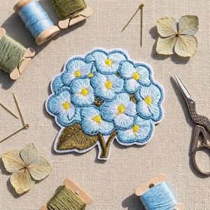 May include: An embroidered patch featuring a cluster of light blue hydrangea flowers with yellow centers and a single dark green leaf. The patch is surrounded by sewing supplies including wooden spools of thread, metal needles, and scissors.