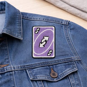 May include: A rectangular patch featuring a purple card with a white border. The card has a central design of two opposing arrows and smaller arrow symbols at the top and bottom. The patch is affixed to a denim jacket.
