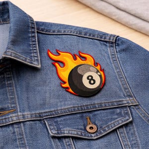 May include: A denim jacket with a patch featuring a black eight ball engulfed in flames. The patch has a yellow and orange flame design with black and white details. The jacket has a classic design with pockets and a button closure.