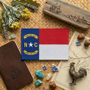 May include: A rectangular patch featuring the North Carolina state flag, with a blue field and the text "MAY 20th 1775" and "APRIL 12th 1776". The flag is red and white. The image includes a wooden carving of a ship, a bronze rooster, and a map.