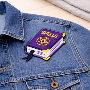 May include: A purple and yellow embroidered patch in the shape of a book with the word "SPELLS" on the cover. The patch features a gold pentagram and is placed on a blue denim jacket.