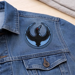 May include: A round, embroidered patch featuring a black bird with outstretched wings and a red eye, set against a light blue circle. The patch is sewn onto a blue denim jacket, with a buttoned pocket visible.