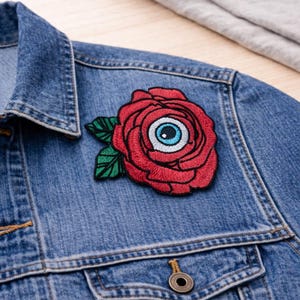 May include: Embroidered patch featuring a red rose with an eye in the center, green leaves, and black outlines. The patch is affixed to a blue denim jacket. The jacket has a buttoned pocket and a button closure.