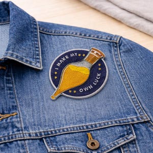 May include: A denim jacket with a circular patch that reads "I MAKE MY OWN LUCK." The patch features a yellow potion bottle design with brown and gold accents, set against a navy blue background with gold stars.