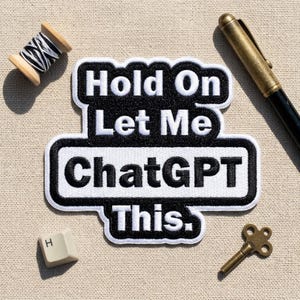 May include: A black and white embroidered patch featuring the text "Hold On Let Me ChatGPT This." The patch is surrounded by a wooden spool of black and white thread, a metallic pen, a single computer keyboard key with the letter H, and a small key.