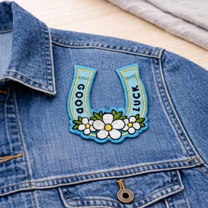 May include: A denim jacket with a decorative patch. The patch is a light blue horseshoe shape with the words "GOOD" and "LUCK" in gold lettering. The horseshoe is decorated with white flowers with yellow centers and green leaves.