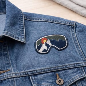 May include: A blue denim jacket with a mountain-themed patch. The patch features a stylized mountain range, a setting sun, and a white mountain peak. The jacket has a classic collar, button closure, and two front pockets.