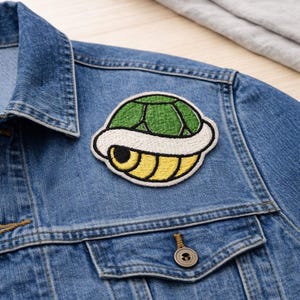 May include: Embroidered patch featuring a green and white Koopa shell design. The patch is sewn onto a blue denim jacket. The shell has a white border and yellow accents. The jacket has a button closure and two chest pockets.