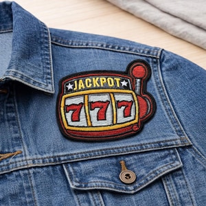 May include: A denim jacket with a colorful embroidered patch. The patch features a slot machine design with the word "JACKPOT" and the number "777". The patch has red, yellow, and black accents.