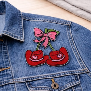 May include: A red embroidered cherry patch with a pink bow and green leaves, featuring a heart detail on each cherry. The patch is affixed to a blue denim jacket, showcasing a playful, decorative accessory.