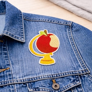 May include: Embroidered patch featuring a red apple globe design with a bite taken out of it. The patch has yellow and green accents and is sewn onto a denim jacket. The patch is a fun accessory for backpacks, jackets, or other fabric items.