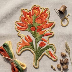 May include: An embroidered patch featuring a vibrant orange and green floral design with yellow accents. The patch is surrounded by embroidery supplies including a brown thimble, two silver needles, a coil of yellow thread, and bundles of embroidery floss.
