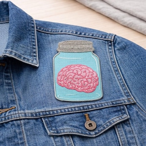 May include: A denim jacket with a patch featuring a pink brain inside a blue jar. The patch has a light blue border and a gray lid. The jacket has a buttoned pocket and a button closure.