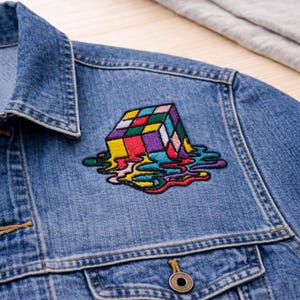 May include: A denim jacket with a colorful embroidered patch. The patch depicts a Rubik's Cube design with a melting effect, featuring vibrant colors like red, yellow, green, and purple. The jacket has a classic button-down design.