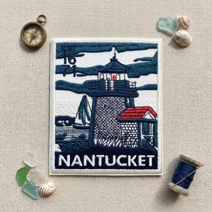 May include: A rectangular embroidered patch featuring a lighthouse scene with a sailboat, dark blue clouds, and a red-roofed structure. The text EST 1671 appears in the upper left, and the word NANTUCKET is embroidered in white at the bottom.