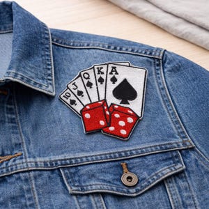 May include: A denim jacket with a patch featuring playing cards and dice. The patch shows a ten, queen, king, and ace of spades, along with two red dice with white dots. The jacket is a light blue wash.