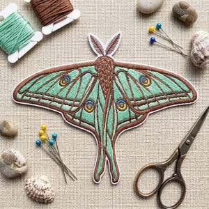 May include: An embroidered moth patch featuring mint green wings with brown borders and detailed brown antennae. The wings display circular eye-like markings in yellow, blue, and brown. The patch is surrounded by sewing supplies and small stones.