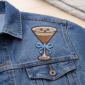 May include: Embroidered patch featuring a brown cocktail glass with coffee beans, a light blue bow, and a cream-colored rim. The patch is affixed to a denim jacket, showcasing its design and potential placement on clothing.