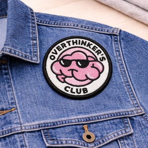 May include: A round patch with the text "OVERTHINKER'S CLUB" encircling a pink brain wearing sunglasses. The patch is on a blue denim jacket.