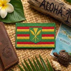 May include: Embroidered patch featuring the Hawaiian flag design with green, red, and yellow stripes and a central emblem. The patch is rectangular and has a detailed, stitched appearance. The patch is on a woven surface with other Hawaiian-themed items.