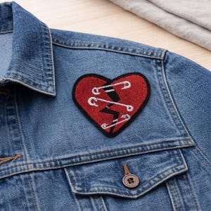 May include: A blue denim jacket with a red heart-shaped patch. The patch features a white stitched design and safety pins, with a black lightning bolt. The jacket has a button closure and a pocket.