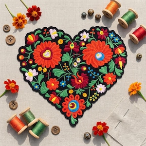 May include: A heart-shaped black fabric patch featuring vibrant floral embroidery in shades of red, orange, yellow, blue, and green. The patch is surrounded by wooden spools of colored thread, loose wooden buttons, and fresh orange flowers.