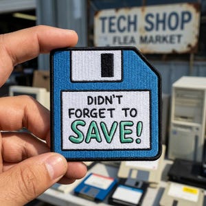 May include: A square embroidered patch shaped like a blue floppy disk with a white label area. The text reads DIDN'T FORGET TO SAVE! in black and green lettering. The patch features a black border and a white sliding tab detail at the top.