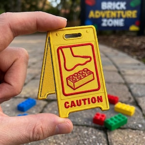 May include: Yellow embroidered patch in the shape of a caution sign, featuring a red outline of a foot stepping on a Lego brick. The word "CAUTION" is in red. The background shows colorful Lego bricks and the text "BRICK ADVENTURE ZONE".