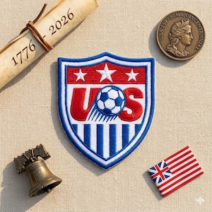 May include: A shield-shaped embroidered patch featuring the red, white, and blue US soccer crest with a central soccer ball. The patch is displayed alongside a rolled scroll marked 1776-2026, a bronze USA 250 semiquincentennial coin, a miniature Liberty Bell, and a small flag.