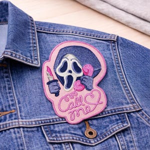 May include: A denim jacket with a decorative patch. The patch features a ghost face design holding a knife and two hearts, with the words "Call me" in pink script. The patch is outlined in pink and blue.