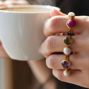 Può includere: Una persona tiene in mano una tazza di caffè bianca con vapore che sale. La persona indossa diversi anelli d'oro con pietre preziose e perle colorate. Gli anelli presentano una varietà di colori, tra cui rosso, marrone, viola e bianco.