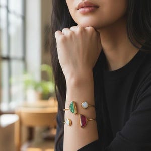 May include: Gold-toned bracelets with colorful gemstone accents. One bracelet features a green stone, another a white stone, and a third a red stone. The bracelets are worn on a person's arm, showcasing the jewelry's design.