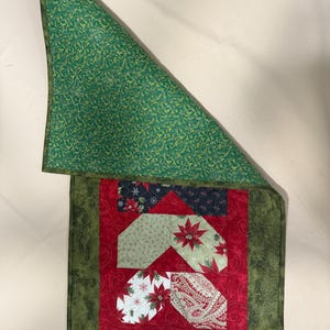 May include: A quilted Christmas table runner with a green border and backing. The centre features red fabric with appliqued shapes in various patterns, including poinsettias and paisley. The runner is approximately 76 cm long.
