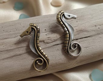 May include: A pair of seahorse earrings. The earrings are made of metal with a two-tone finish, featuring a gold-tone head and body, and a silver-tone body. The seahorses are placed on a piece of driftwood, with seashells and sea glass scattered around.