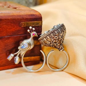 Könnte beinhalten: Ein silberner Ring mit einem Pfauenmotiv und einem dekorativen floralen Muster. Der Ring zeigt einen Pfau mit rotem Auge und blauen Akzenten sowie ein kegelförmiges Element mit aufwendigen Details. Der Ring wird auf einer beigen Oberfläche präsentiert.