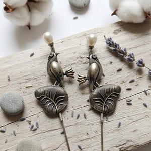 May include: A pair of silver-coloured bird hat pins, each with a pearl at the top, resting on leaf-shaped bases. The pins are arranged on a wooden board with cotton, small stones, and lavender, creating a natural, rustic display.