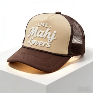 Puede incluir: Gorra de camionero marrón y beige con el texto blanco "-LME- Mahj Lovers" y un corazón. La gorra tiene una visera marrón y una parte trasera de malla. Botón en la parte superior.