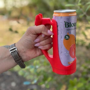 May include: A red 3D-printed can holder with a handle, holding a can of Bloom sparkling water. The can features a peach and mango graphic, with the text "Peach Mango" and "0g Sugar".