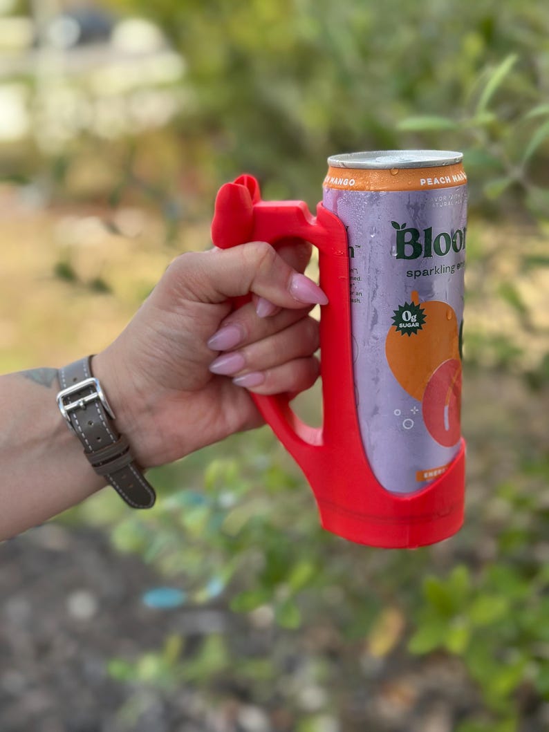 May include: A red 3D-printed can holder with a handle, holding a can of Bloom sparkling water. The can features a peach and mango graphic, with the text "Peach Mango" and "0g Sugar".