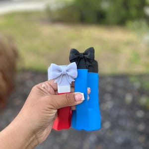 May include: Three colorful bottle stoppers with bow accents. The stoppers are red, blue, and black, each topped with a bow in a matching or complementary color. The stoppers are held in a hand against a blurred outdoor background.