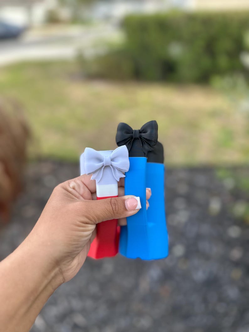 May include: Three colorful bottle stoppers with bow accents. The stoppers are red, blue, and black, each topped with a bow in a matching or complementary color. The stoppers are held in a hand against a blurred outdoor background.