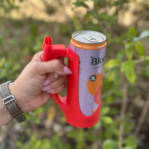 May include: A red 3D-printed can holder with a handle, holding a can of Bloom sparkling water. The can is purple with orange accents and the words "Peach Mango" and "Bloom" are visible. The background is blurred green foliage.